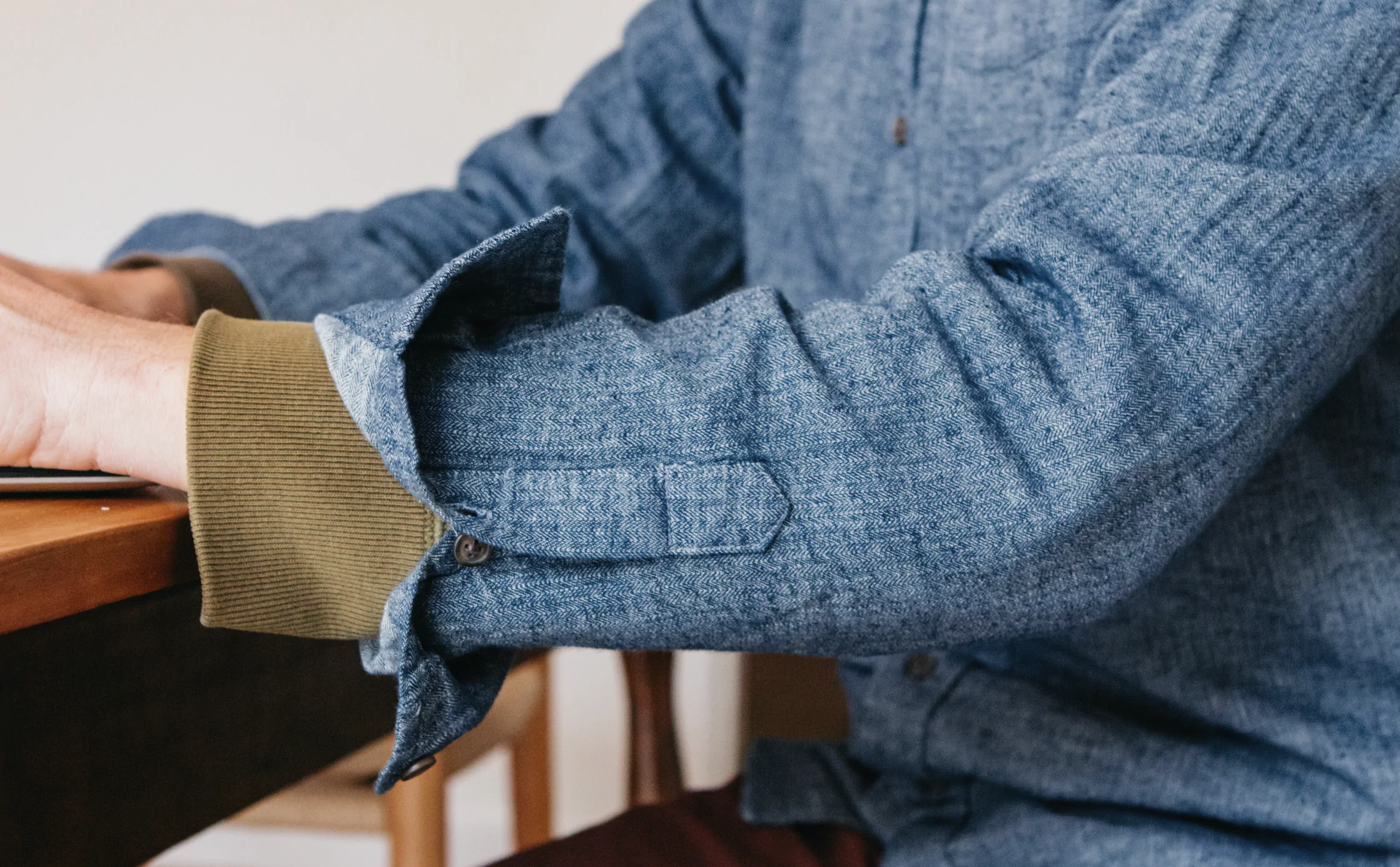 A person wearing a blue button-up shirt with rolled-up sleeves and a green sweater cuff sits at a table, using a laptop or computer, only their arm and torso visible.