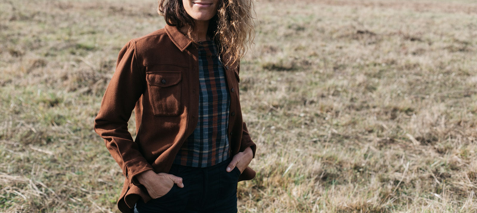 A person stands in a grassy field wearing a brown jacket over a plaid shirt with hands in pockets. The photo is cropped at the mouth, so their full face isn’t visible.