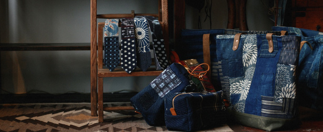 A wooden chair displays patterned neckties in blue tones, with matching blue bags and totes featuring patchwork designs arranged nearby on a geometric rug.