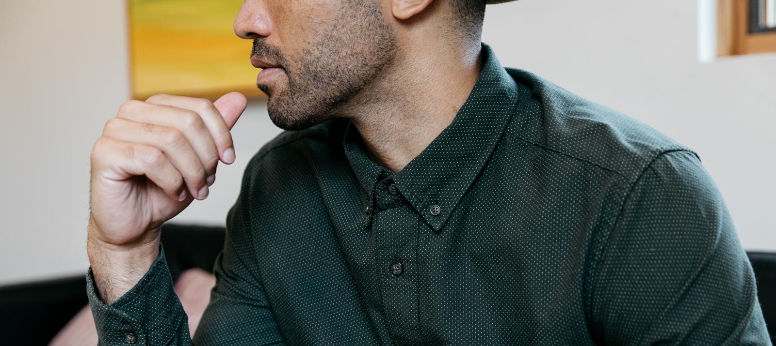 A man wearing a dark green, buttoned-up shirt sits indoors, resting his chin on his hand. His face is partially visible, and the background is softly blurred.