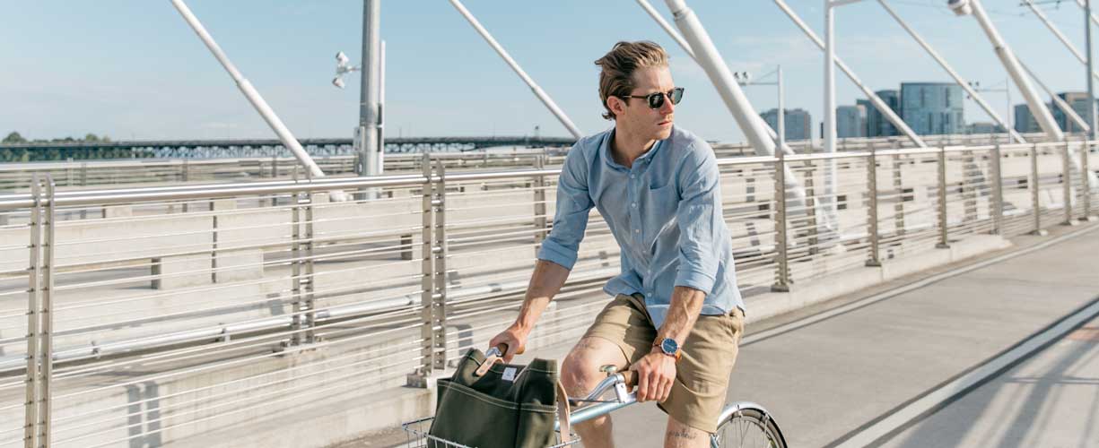 A man wearing sunglasses, a light blue shirt, and khaki shorts rides a bicycle across a modern pedestrian bridge with metal railings on a sunny day.