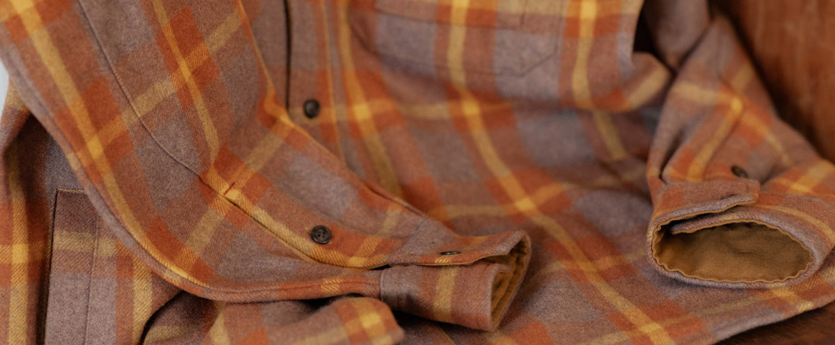 A close-up of a soft, brown and orange plaid flannel shirt with buttoned cuffs and a chest pocket, lying casually folded on a wooden surface.