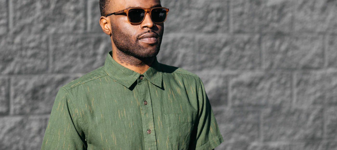 A man wearing sunglasses and a green short-sleeve button-up shirt stands in front of a gray brick wall, looking confidently ahead.