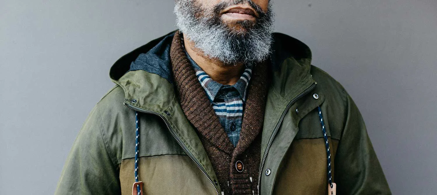 A man with a gray beard wears glasses, a green hooded jacket, a brown sweater, and a collared shirt with blue and white stripes, standing against a plain gray background.