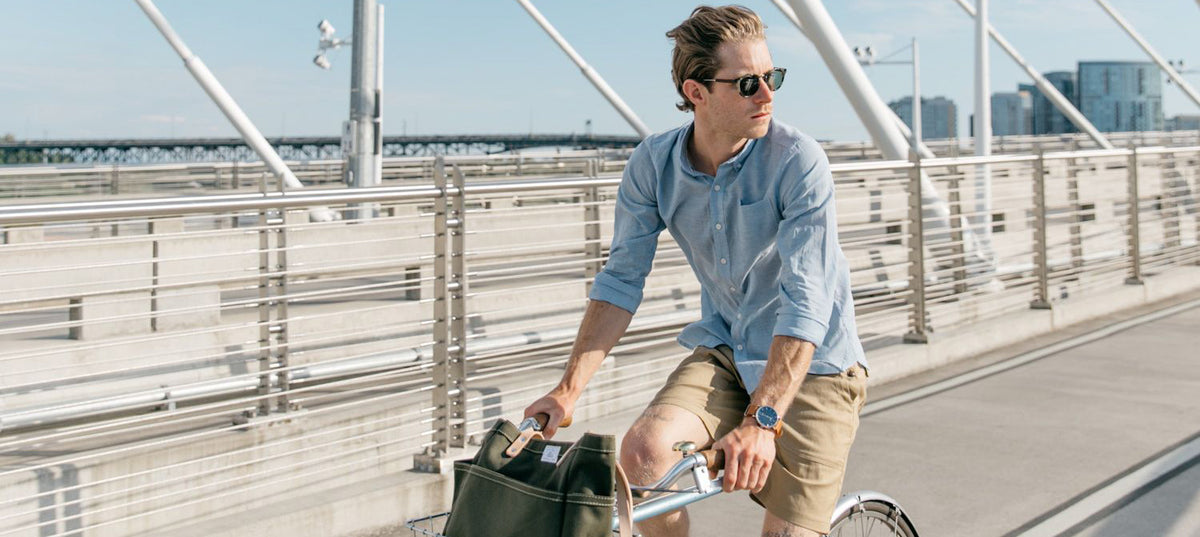 A man wearing sunglasses, a light blue shirt, and khaki shorts rides a bicycle on a modern pedestrian bridge on a sunny day, with city buildings visible in the background.