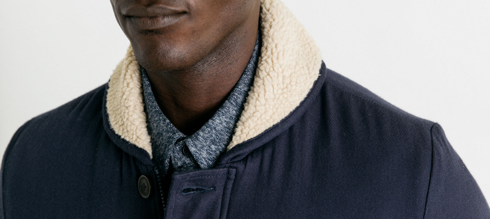 Close-up of a person wearing a navy jacket with a cream sherpa collar over a grey button-up shirt, against a plain light background. The persons face is partially visible.
