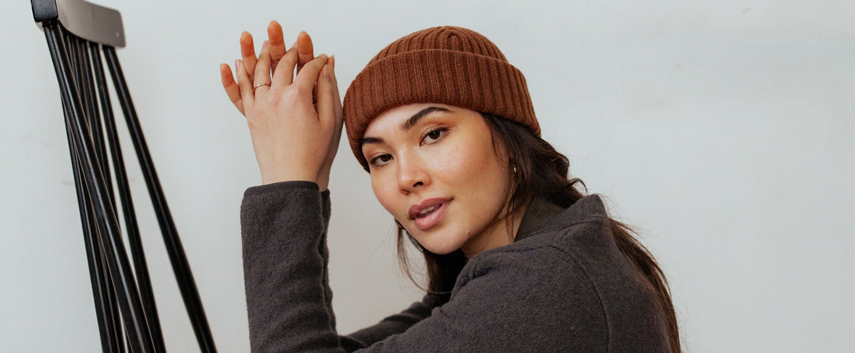 A woman wearing a brown knit beanie and a dark jacket sits beside a black easel, looking at the camera with her hands clasped above her head.