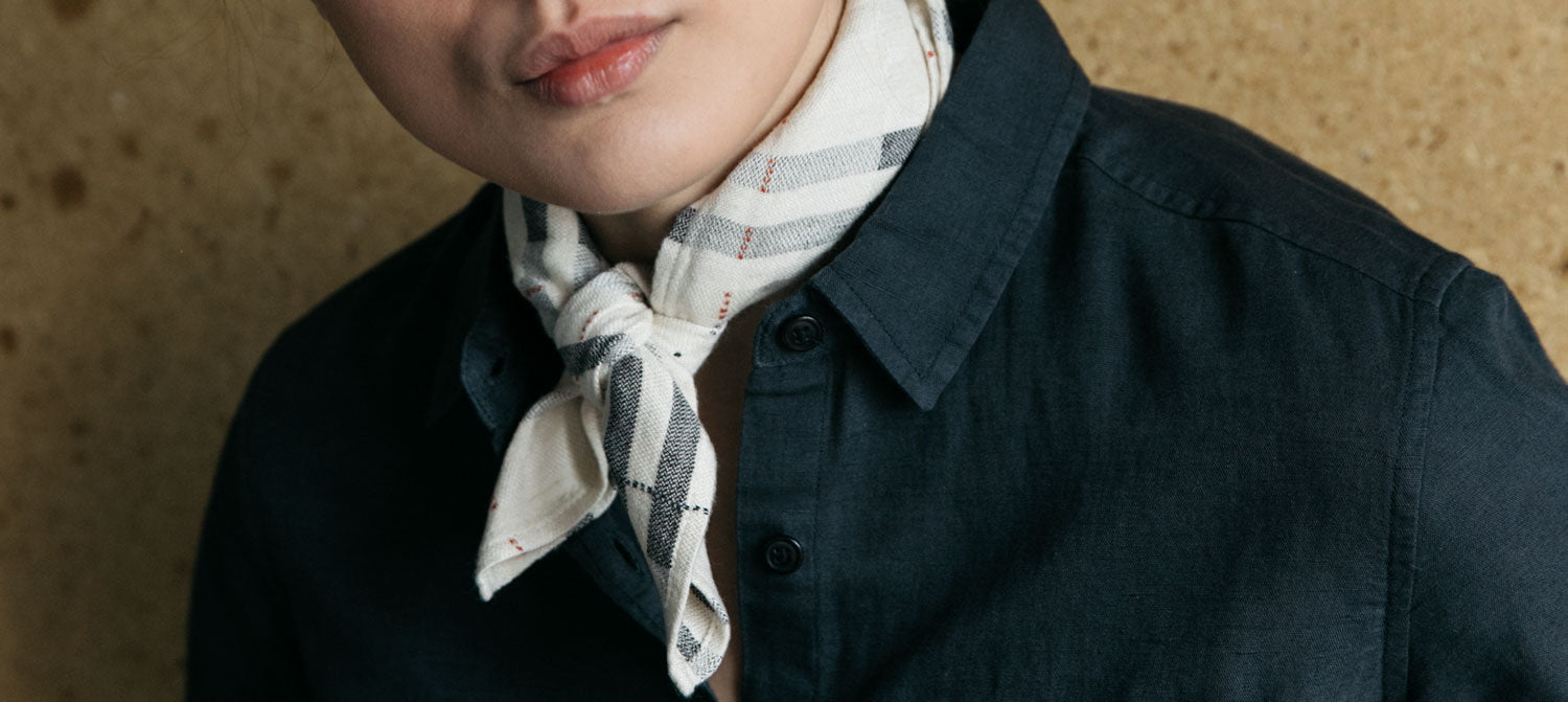 A person wears a dark button-up shirt and a white plaid scarf knotted around their neck, standing against a light brown textured background.