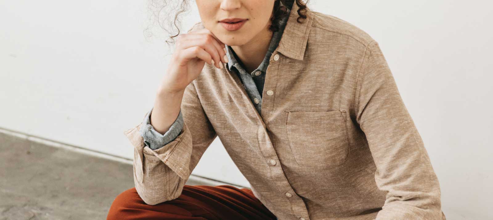A person with curly hair, wearing a beige button-up shirt over a blue collared shirt and rust-colored pants, sits indoors with one hand resting near their chin.