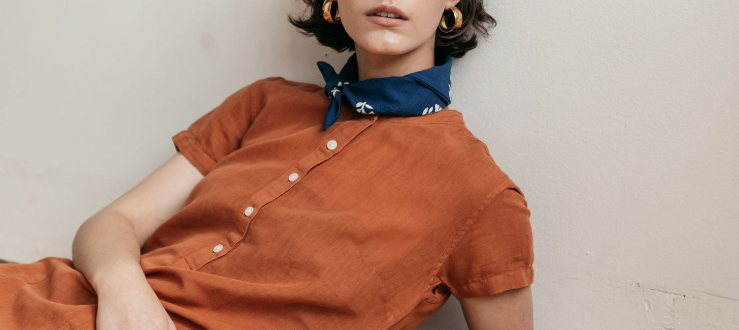 A person with short dark hair wearing gold hoop earrings, a blue bandana around their neck, and an orange short-sleeve button-up dress, sitting against a light-colored wall.