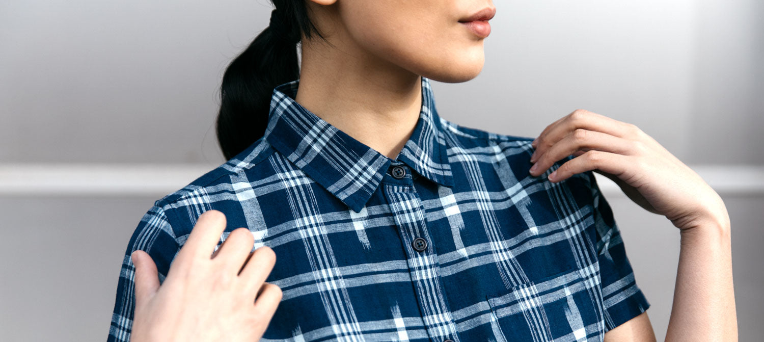 A person with dark hair in a ponytail adjusts the collar of a blue and white plaid shirt, shown from the shoulders up against a neutral background.