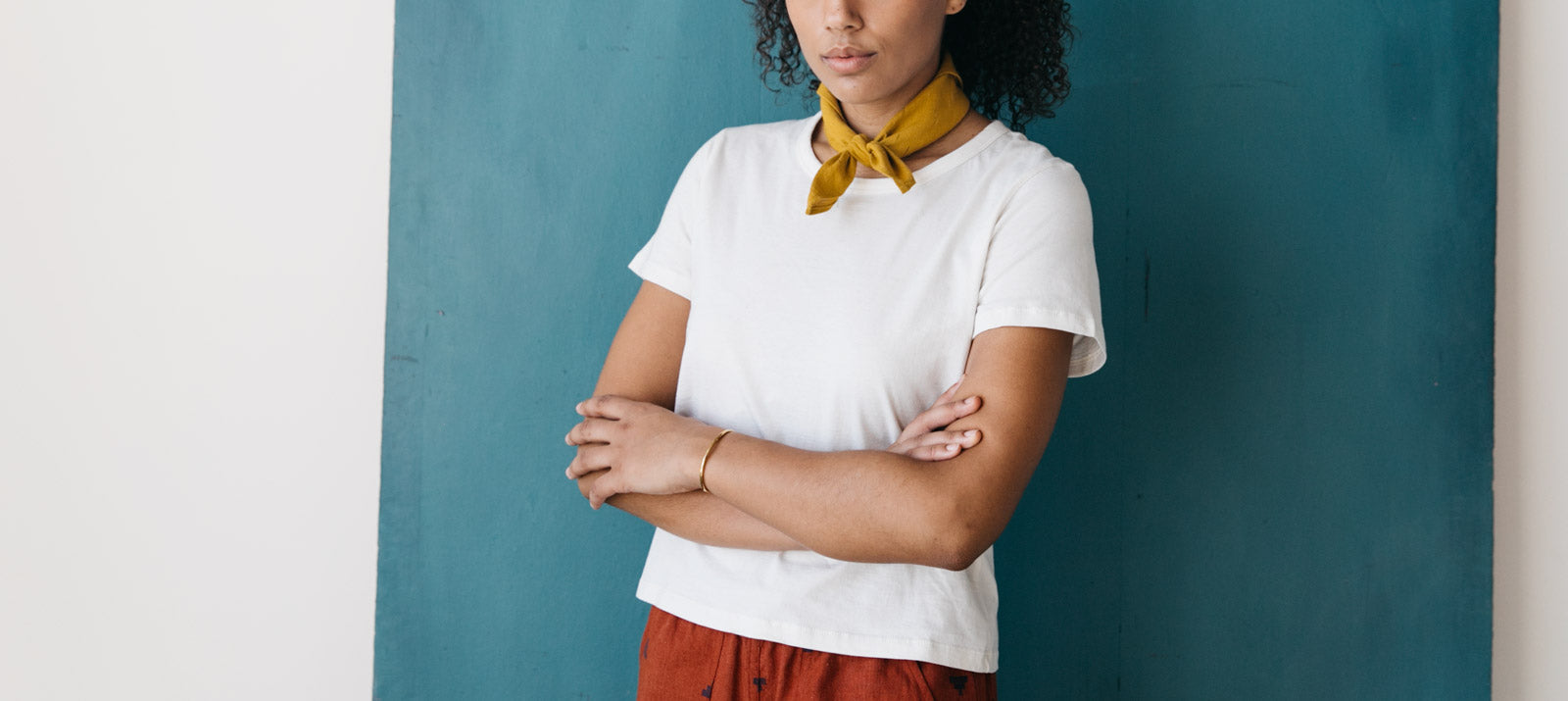 A person stands with arms crossed, wearing a white t-shirt, red pants, and a yellow scarf around their neck, in front of a blue background. Their face is partially out of frame.