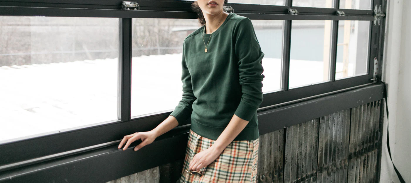 Person in a green sweater and plaid skirt stands by large industrial windows, resting one hand on the windowsill, with soft natural light coming in.