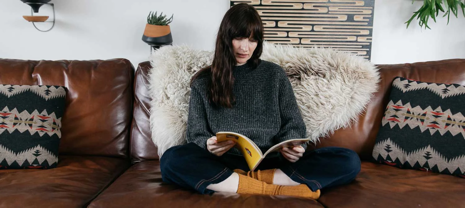 A person with long brown hair sits cross-legged on a brown leather couch, reading a book. Decorative pillows and plants are visible in the cozy, modern living room.