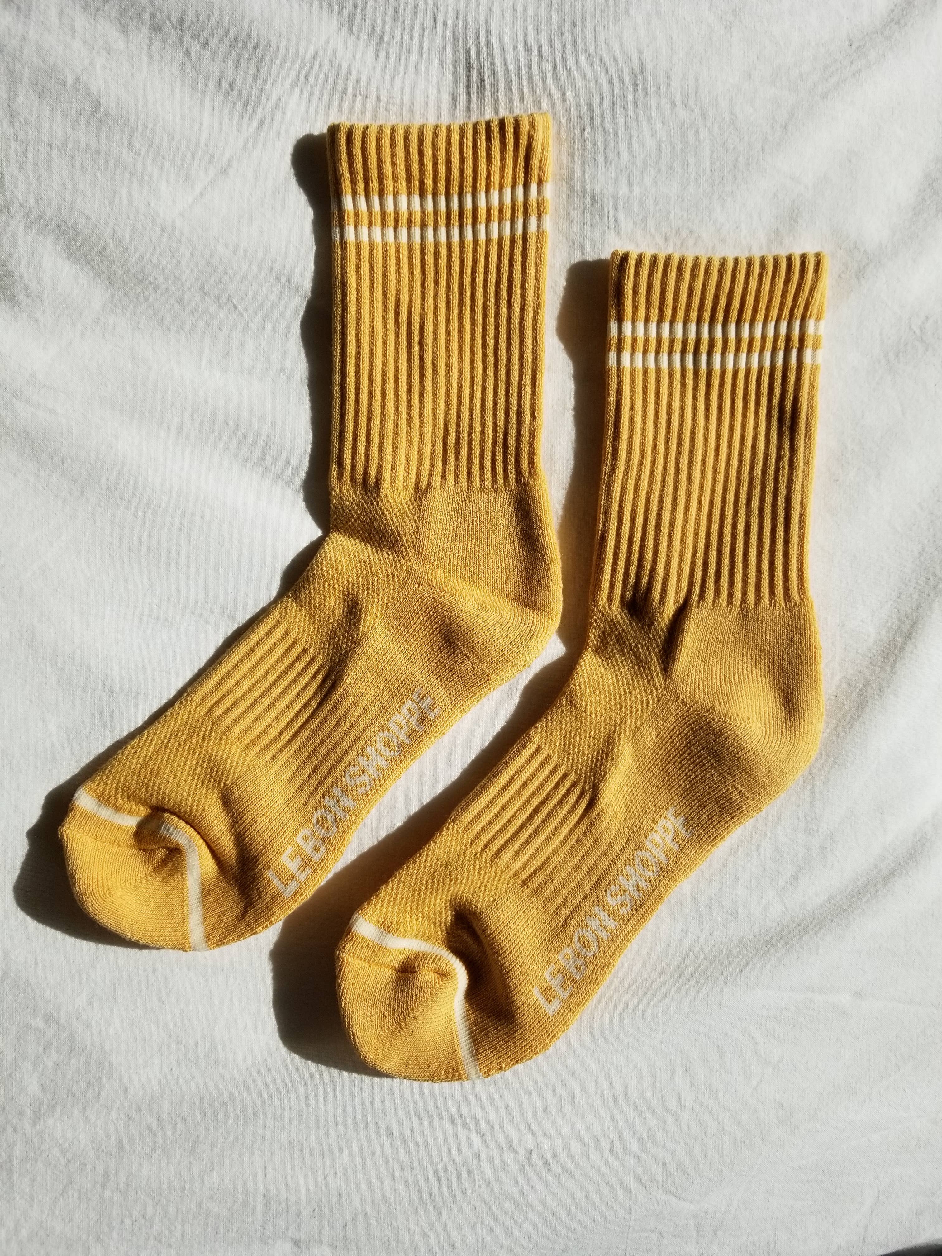 A pair of Le Bon Shoppe Boyfriend Socks in Butter, featuring yellow ribbed fabric with two white stripes near the top and "LEBONSHOPPE" on the soles—ideal for fans of cute, sporty socks.