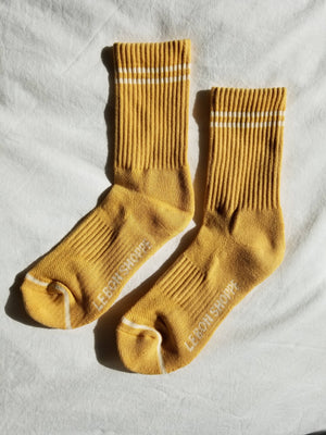 A pair of Le Bon Shoppe Boyfriend Socks in Butter, featuring yellow ribbed fabric with two white stripes near the top and "LEBONSHOPPE" on the soles—ideal for fans of cute, sporty socks.