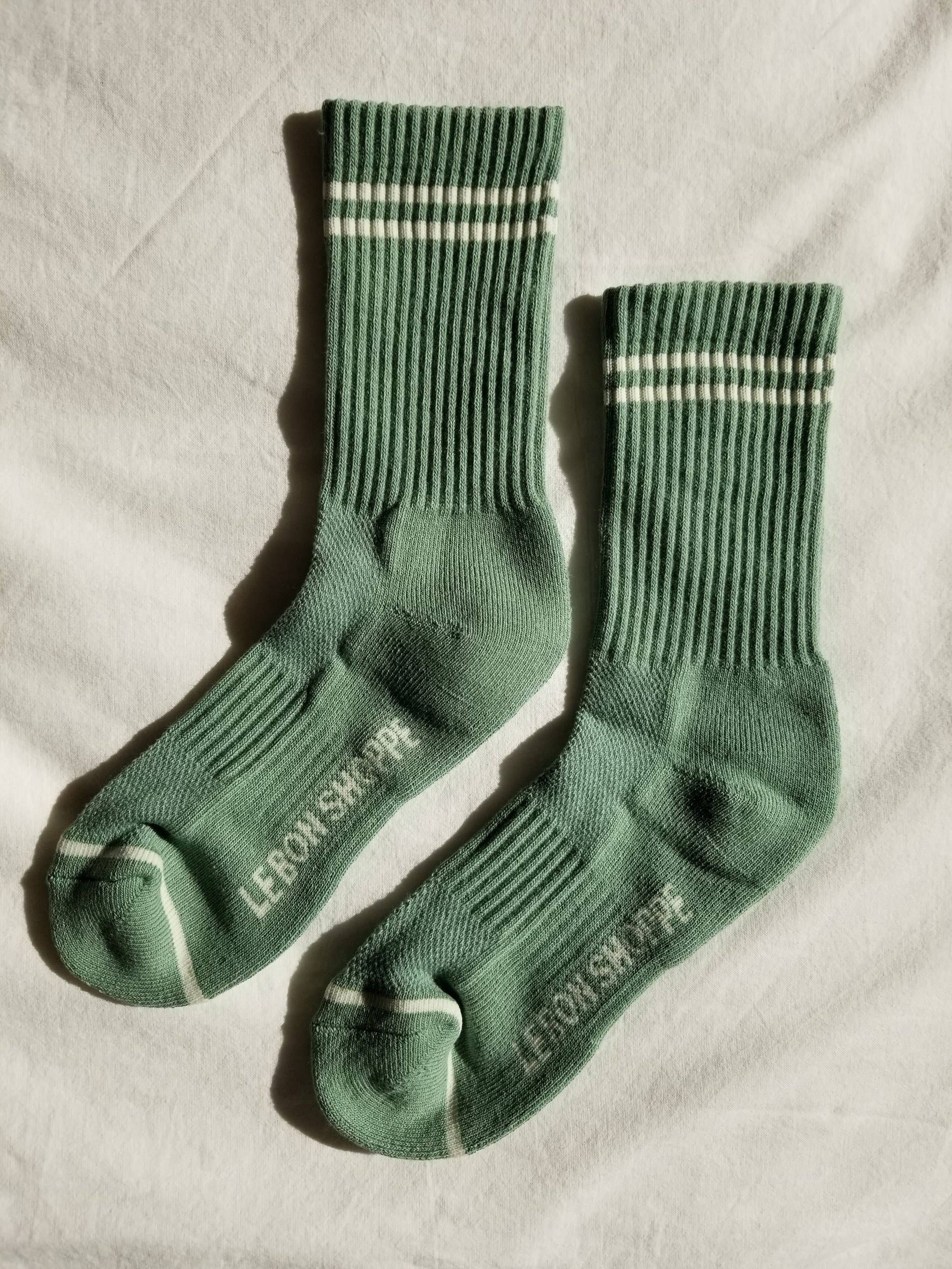 A pair of Le Bon Shoppe - Boyfriend Socks in Meadow, featuring white stripes near the top and "LEBONSHOPPE" text on the soles, are laid out on a white fabric background—these cute socks add a sporty touch to any outfit.