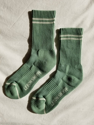 A pair of Le Bon Shoppe - Boyfriend Socks in Meadow, featuring white stripes near the top and "LEBONSHOPPE" text on the soles, are laid out on a white fabric background—these cute socks add a sporty touch to any outfit.