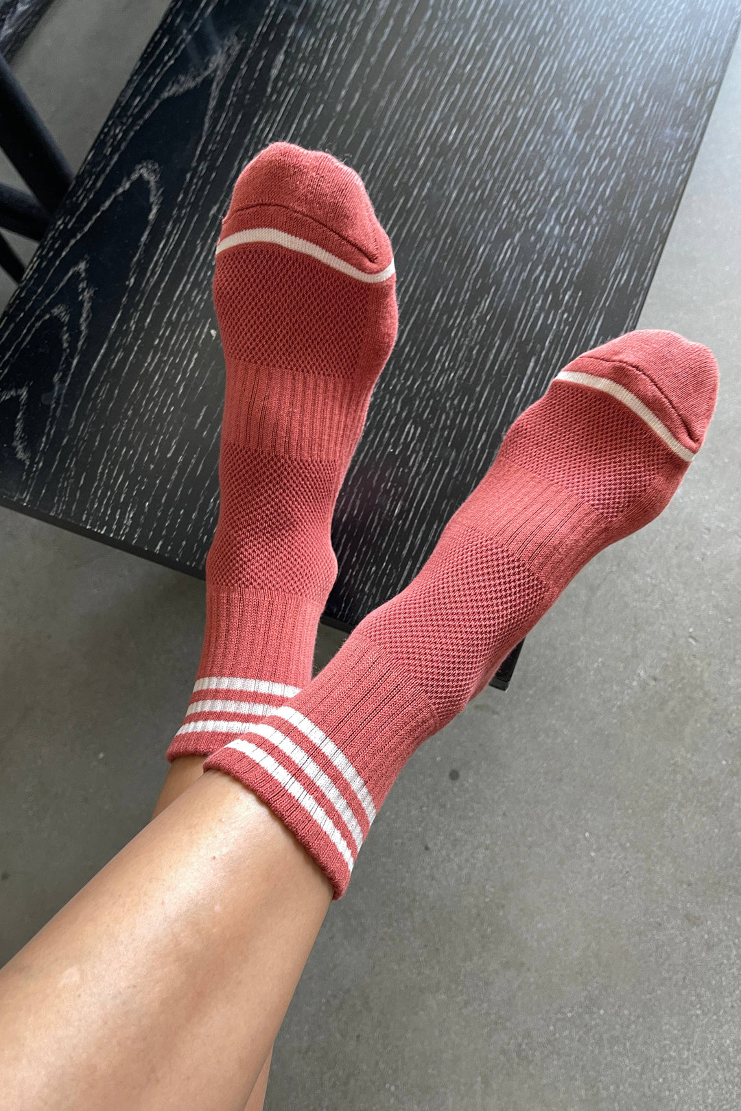 A person wears Le Bon Shoppe - Girlfriend Socks in Terracotta, feet up on a dark wood table with a gray floor in the background. These cozy Le Bon Shoppe socks add a playful touch to any casual moment.