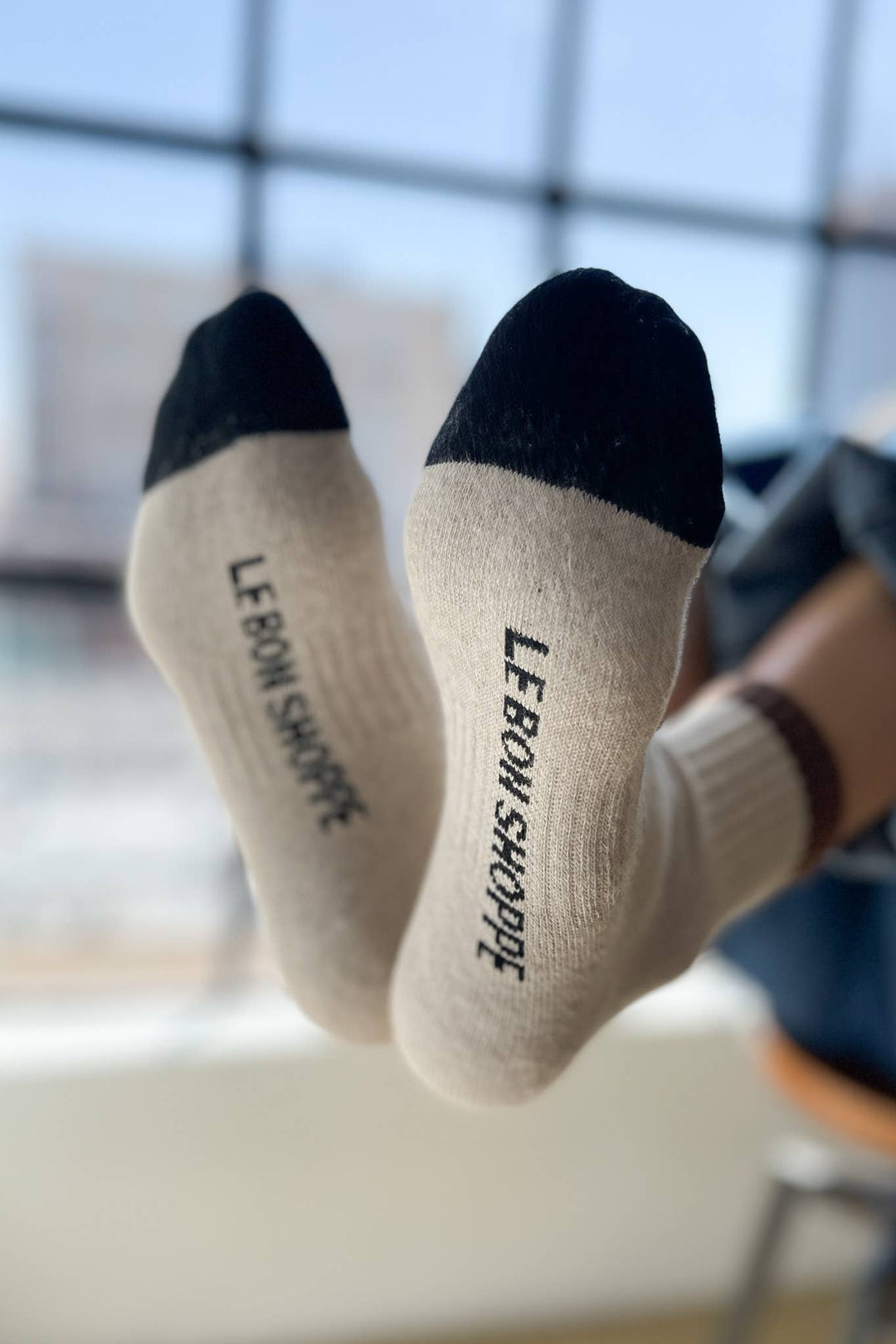 A close-up of two feet wearing Le Bon Shoppe - Color Block Girlfriend Socks in Black Coffee—beige socks with black toes. Soft natural light streams through a window in the blurred background.