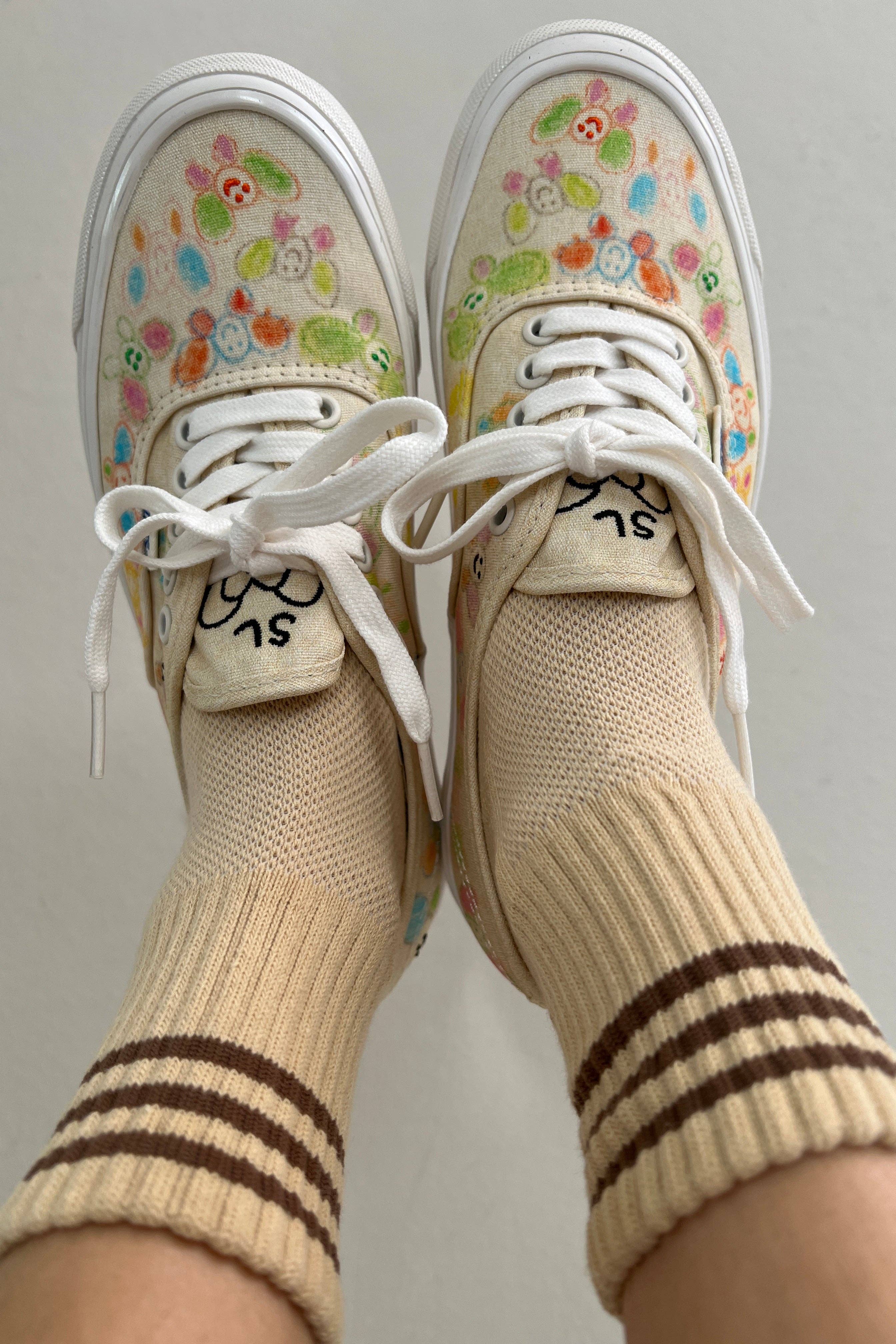 A close-up of feet in Le Bon Shoppe - Girlfriend Socks / Daisy crew socks with brown stripes, paired with white sneakers featuring hand-drawn flowers and smiley faces, against a plain light-colored background.