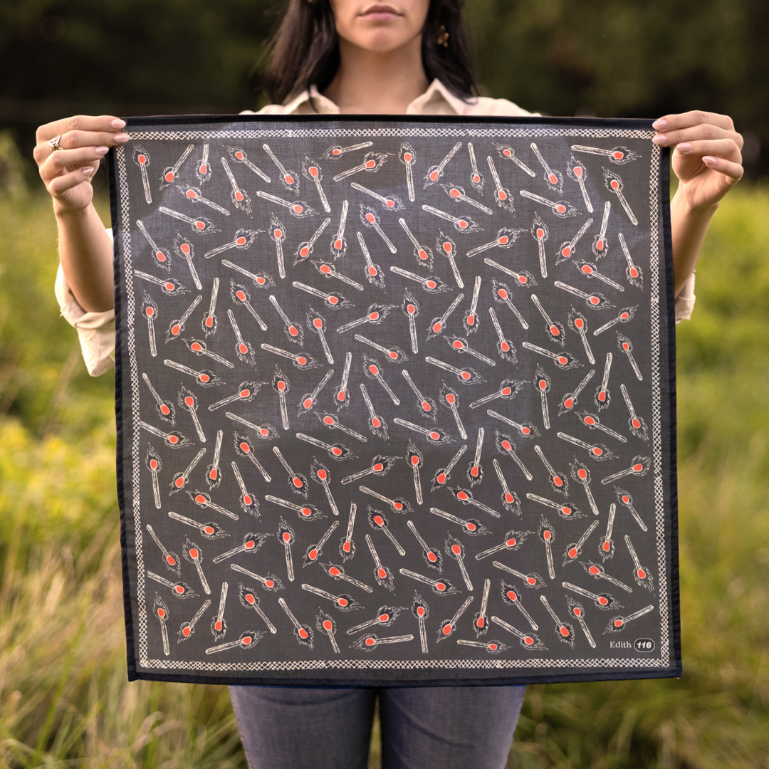 A person stands outdoors holding up the Handker Bandanas - No. 116 Edith Bandana by Handker Bandanas, featuring a charcoal gray color with small red and white spoon illustrations. Grass and greenery blur in the background.