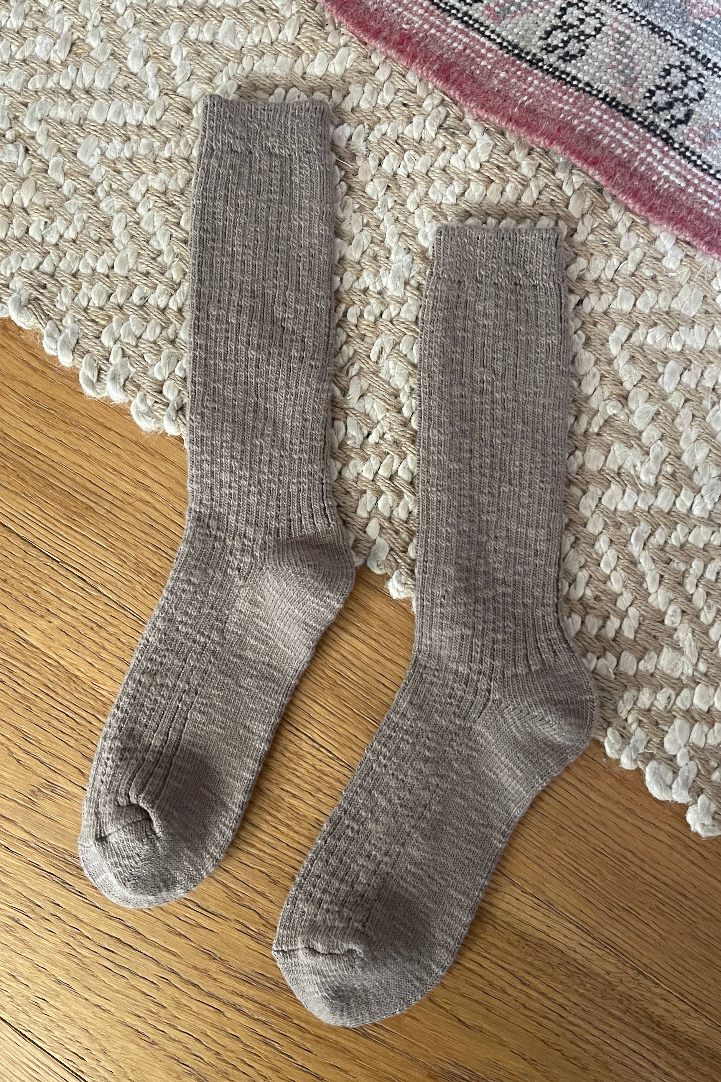 A pair of Le Bon Shoppe Cottage Socks in Flax rests side by side on a textured cream rug and wooden floor, with a pink-patterned rug visible in the corner.