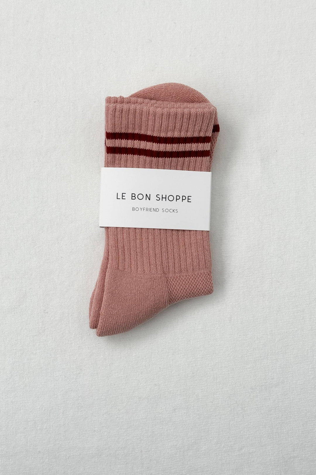 A pair of Le Bon Shoppe - Boyfriend Socks in Vintage Pink, featuring darker pink stripes and a signature white label, folded neatly on a light, textured surface.