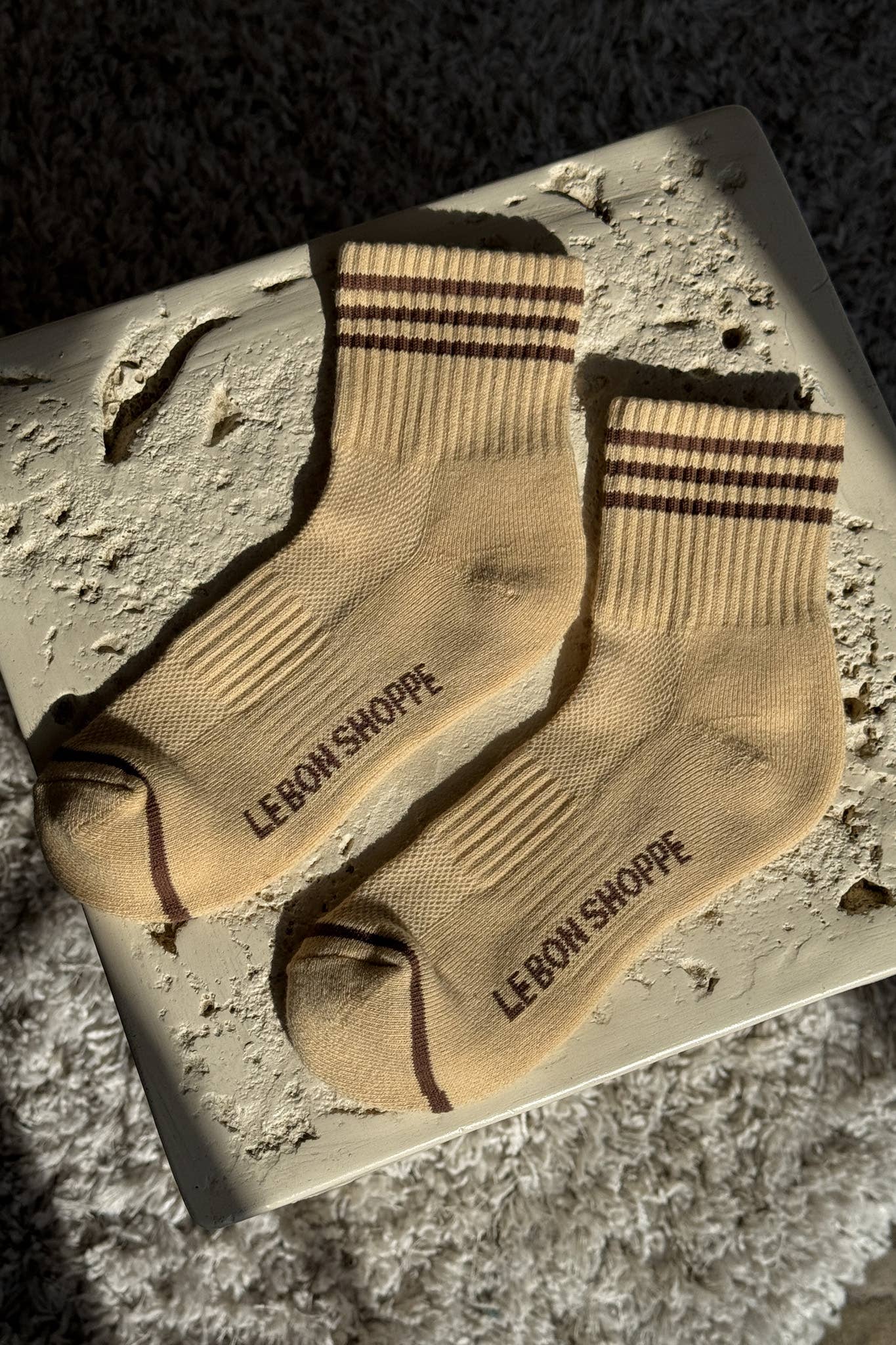 A pair of Le Bon Shoppe - Girlfriend Socks / Daisy in beige with brown stripes and "LE BON SHOPPE" on the side, laid flat on a textured cream surface in sunlight.