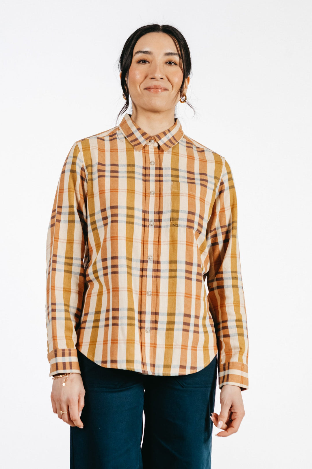 A person with long dark hair wears the Bridge & Burn Ash Slim Shirt in Heceta Plaid, featuring yellow, orange, and brown tones. They stand facing the camera in dark pants against a plain white background and smile gently.