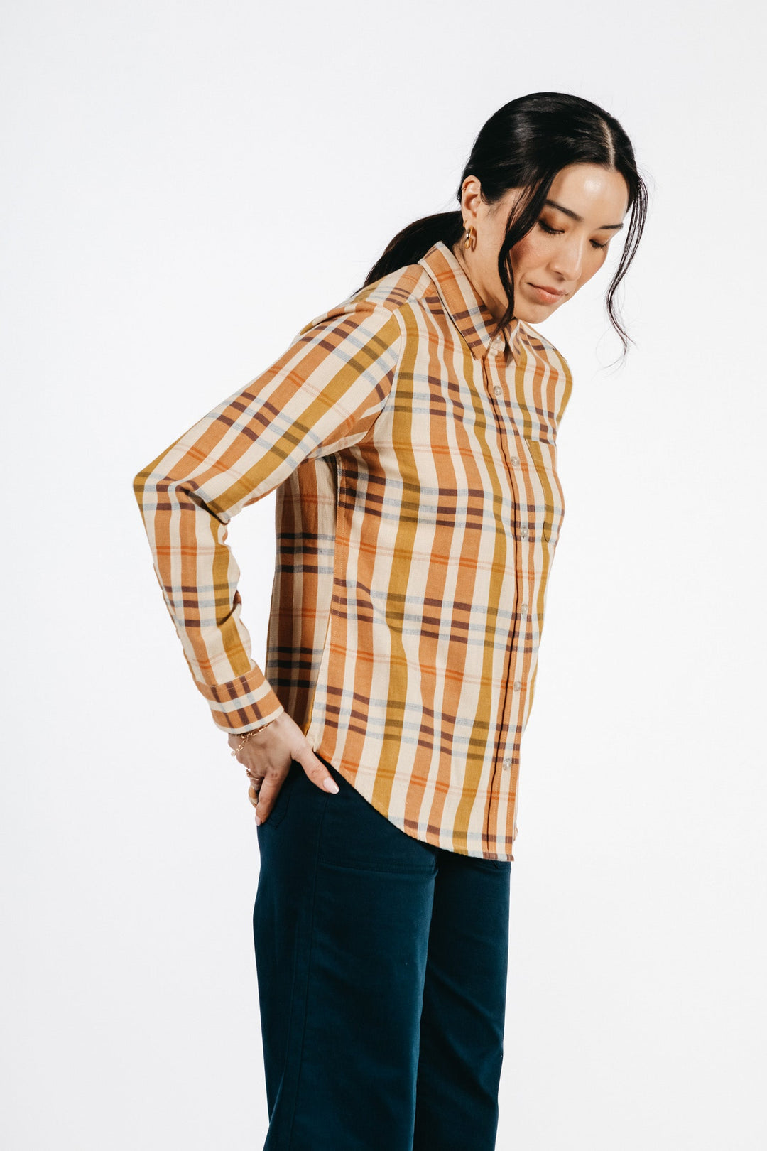 A woman with long dark hair tied back stands sideways against a plain white background, wearing the Bridge & Burn Ash Slim Shirt in Heceta Plaid and dark pants, with one hand resting on her lower back.