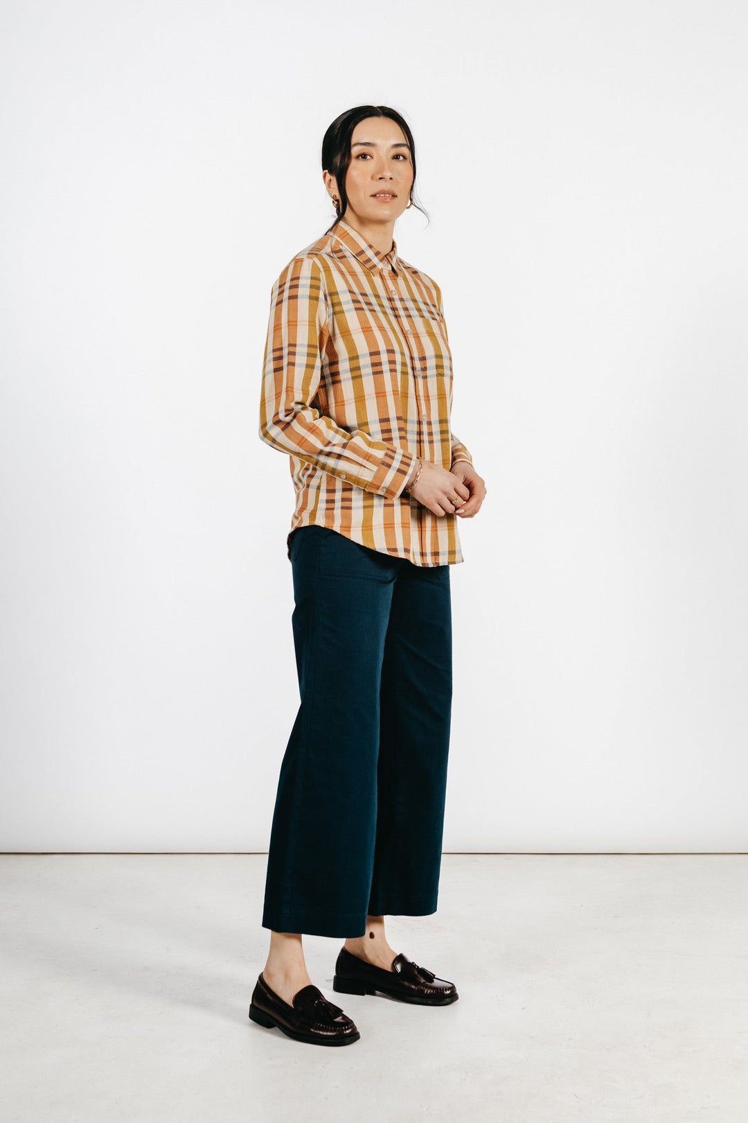 A person with dark hair pulled back stands against a white background, wearing the Bridge & Burn Ash Slim Shirt in Heceta Plaid—yellow and brown plaid—paired with dark wide-leg pants and black loafers, sporting a neutral expression.