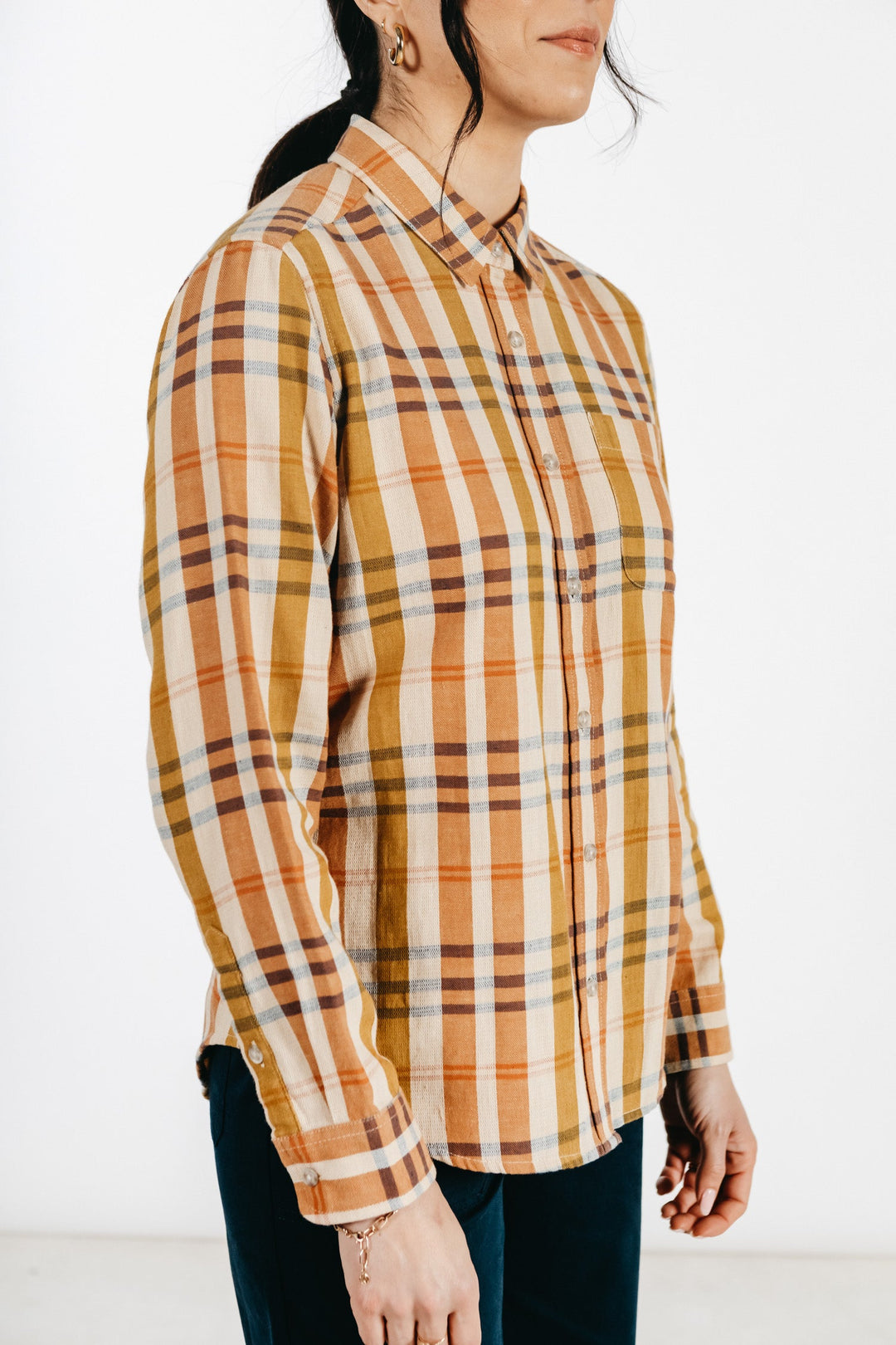 A woman with dark hair in a ponytail wears the Bridge & Burn Ash Slim Shirt in Heceta Plaid—an orange, yellow, and beige button-up—standing against a plain white background.