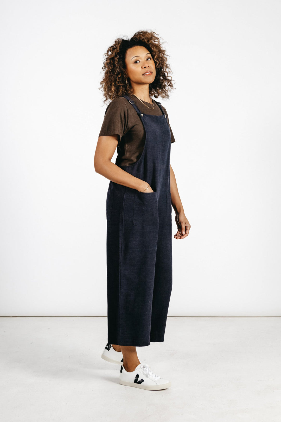 A woman with curly hair stands against a plain white background, one hand in her pocket, wearing the Bridge & Burn Edie Overall in Navy—a wide leg style crafted from a linen-cotton blend—layered over a brown t-shirt and paired with white sneakers.
