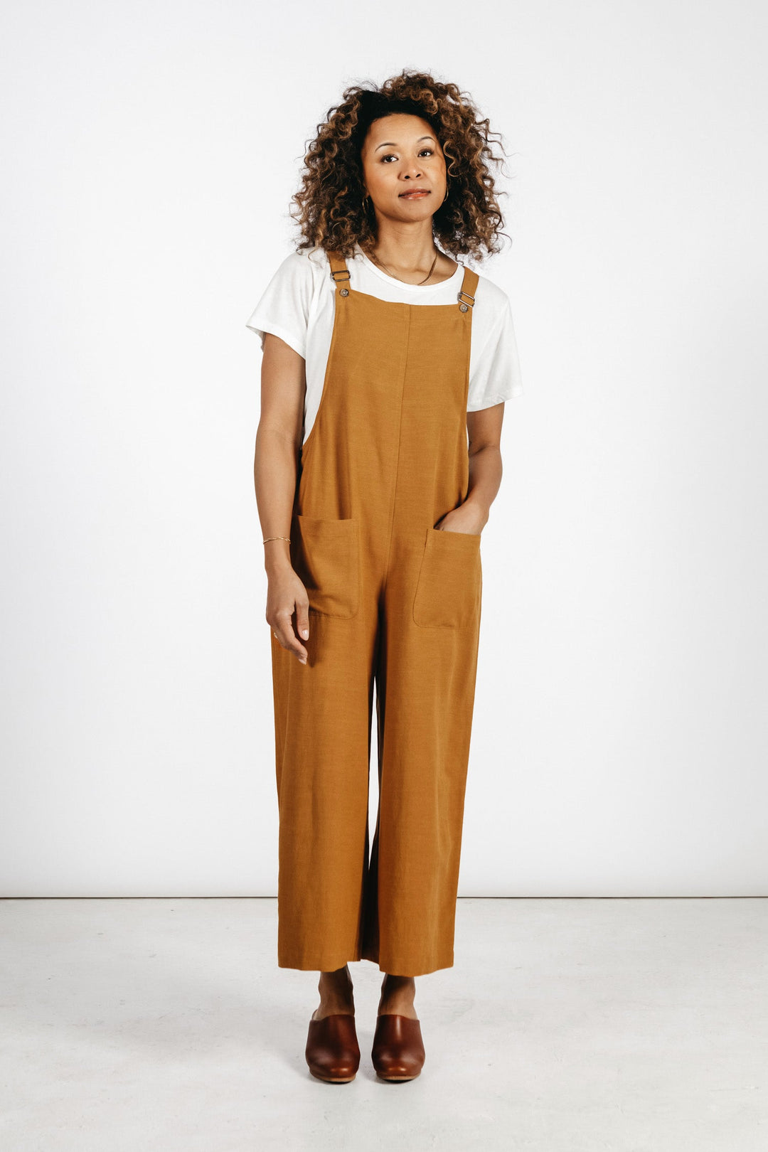 A person with curly hair stands against a white background wearing a white t-shirt, brown shoes, and the Bridge & Burn Edie Overall in Ochre, featuring wide legs, adjustable straps, and one hand in their pocket.