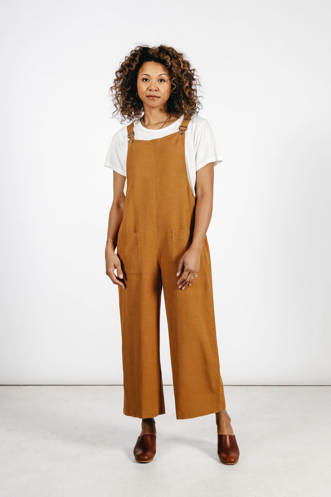 A woman with curly hair stands against a white background, wearing the Bridge & Burn Edie Overall in Ochre over a white t-shirt and brown shoes. She faces forward with a neutral expression.