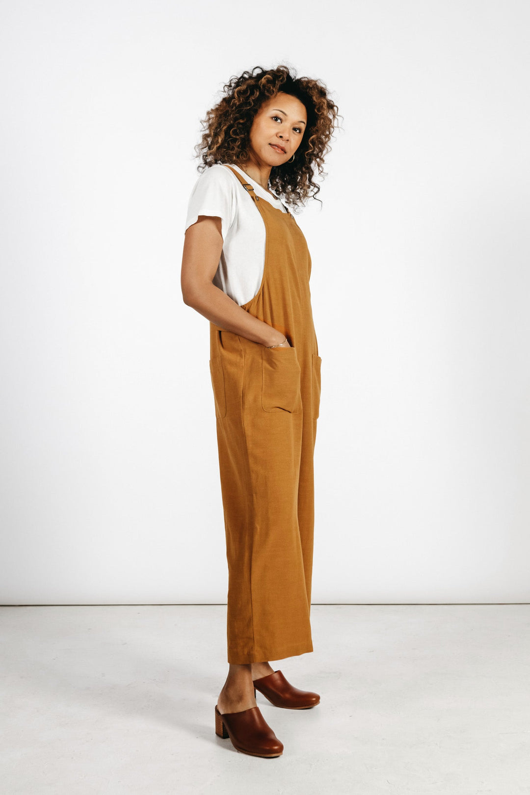 A woman with curly hair wears the Bridge & Burn Edie Overall in Ochre over a white t-shirt, paired with brown heeled shoes, standing against a plain white background.