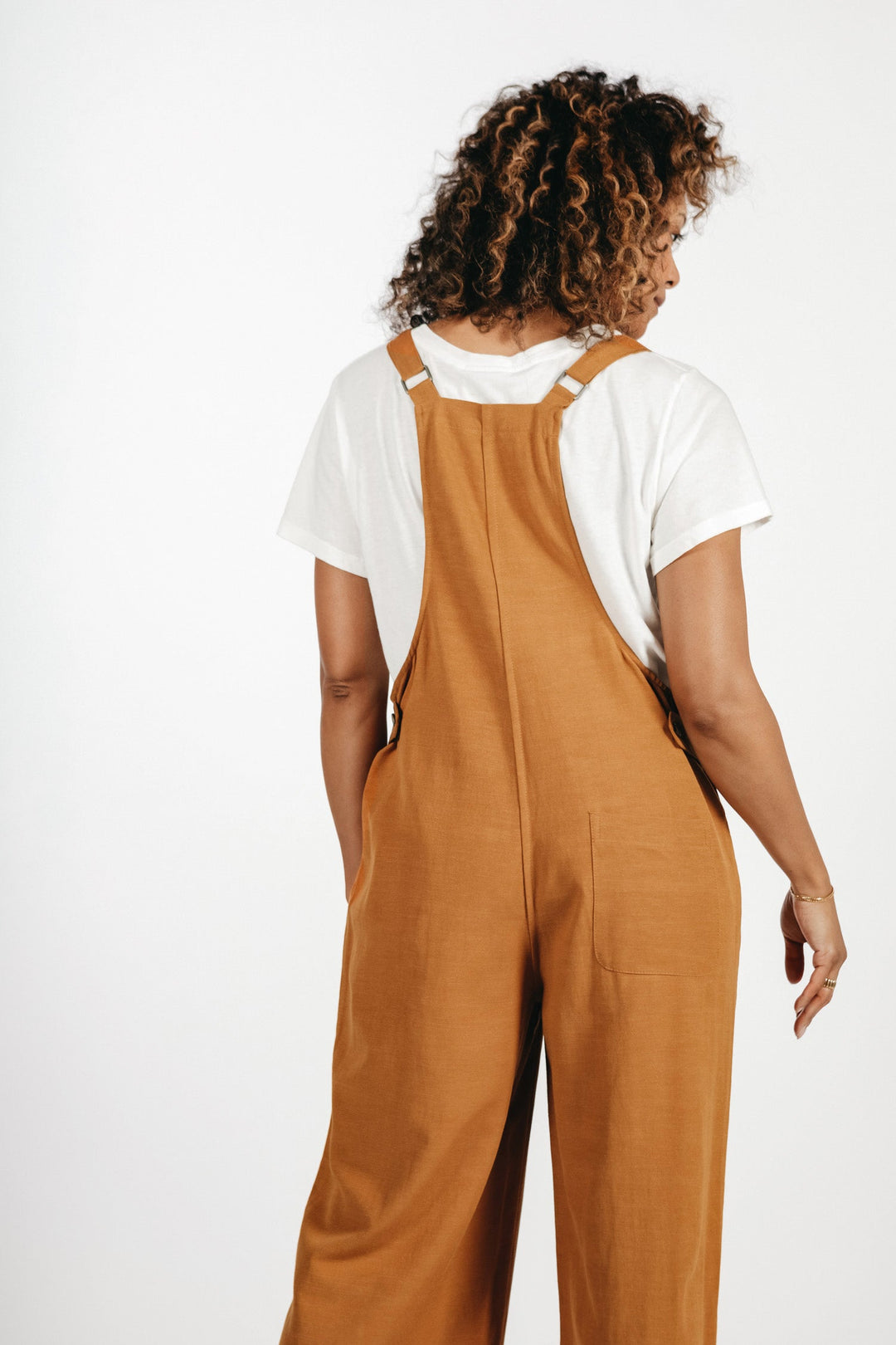 A person with curly hair, seen from behind, wears a white t-shirt and Bridge & Burn’s Edie Overall in Ochre with adjustable straps, standing against a plain white background.