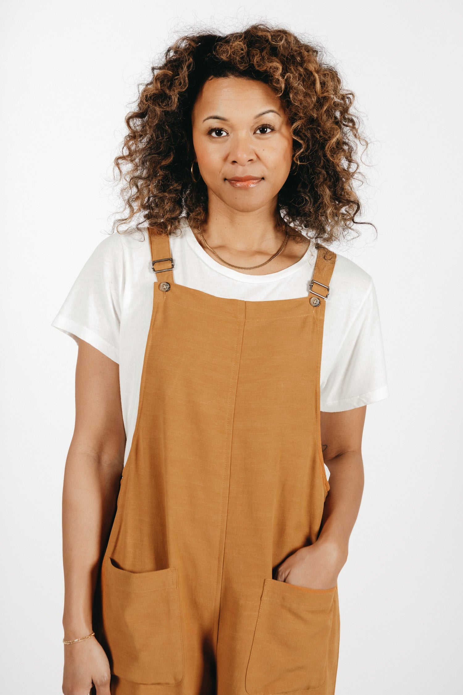 A woman with curly hair stands against a white background, wearing the Bridge & Burn Edie Overall in Ochre over a white linen-cotton tee, one hand in her pocket and a relaxed expression.