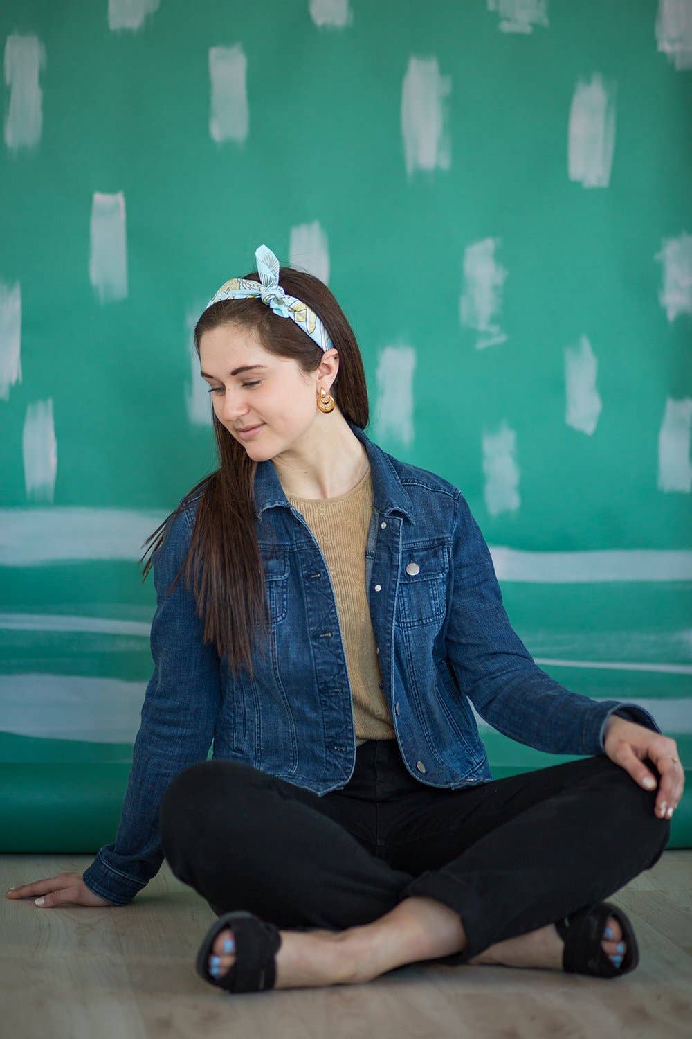 A woman with long brown hair, wearing the No. 012 Eliza Bandana from Handker Bandanas, a denim jacket, beige sweater, and black pants sits cross-legged on a wooden floor against a green-and-white backdrop, smiling as she looks down.