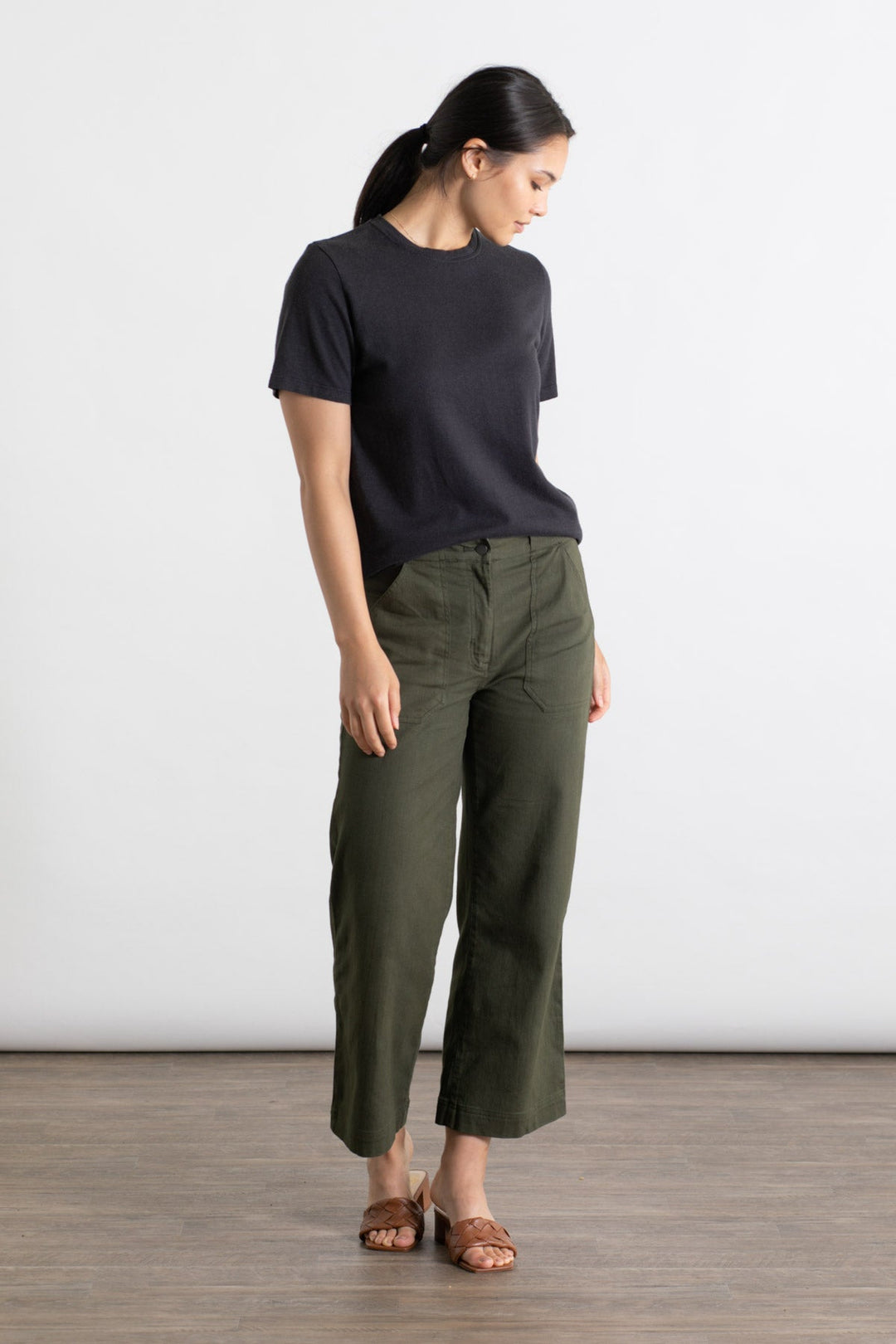 A woman stands indoors, looking down, wearing the Jungmaven Ojai Tee in black with short sleeves, paired with olive green wide-leg pants and brown sandals. Her dark hair is tied back in a ponytail.