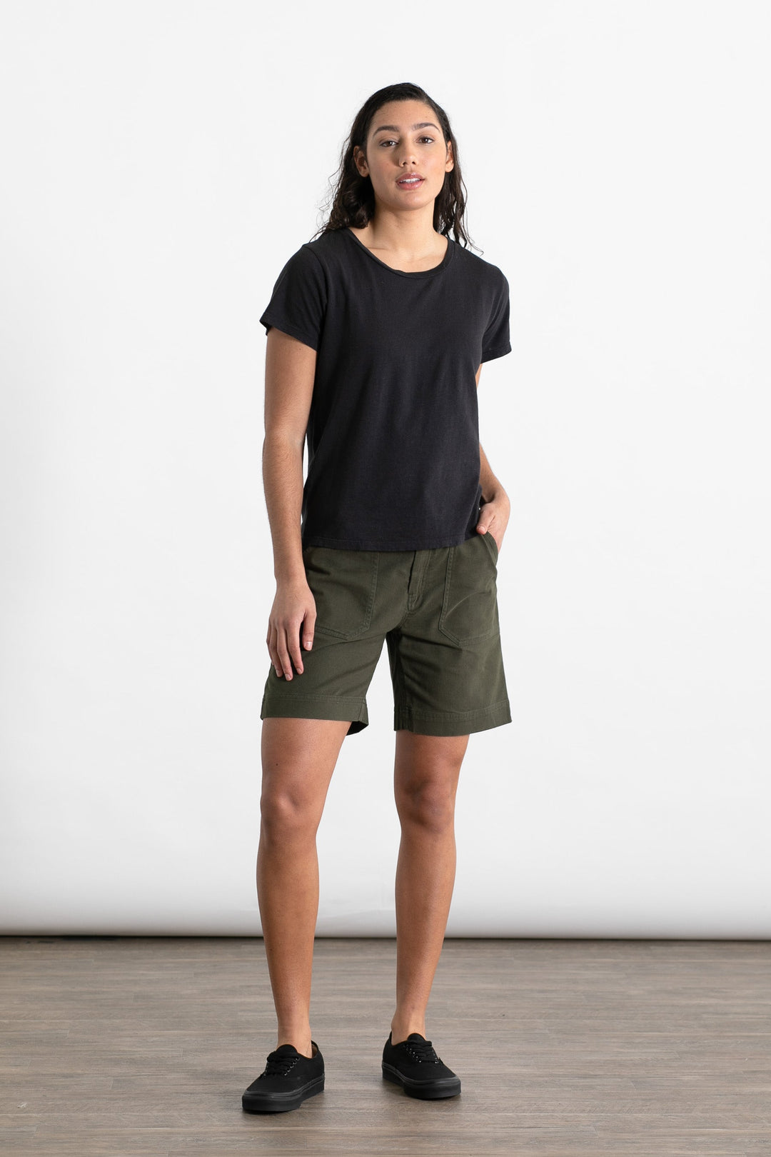 A person stands indoors on a wooden floor wearing the Jungmaven Ojai Tee in black, paired with olive green shorts and black sneakers. With one hand in their pocket, they face the camera against a plain white background.