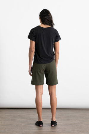 A person with long dark hair, wearing the Jungmaven Ojai Tee in Black, stands facing away from the camera on a wooden floor with a white background.