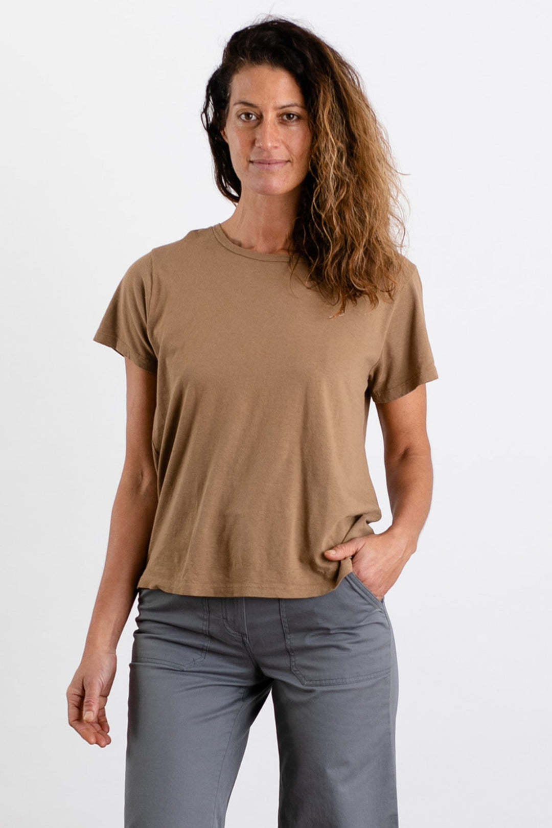 A woman with wavy brown hair wears the Jungmaven Ojai Tee in Coyote, paired with gray pants, standing against a plain white background with one hand in her pocket, looking at the camera.