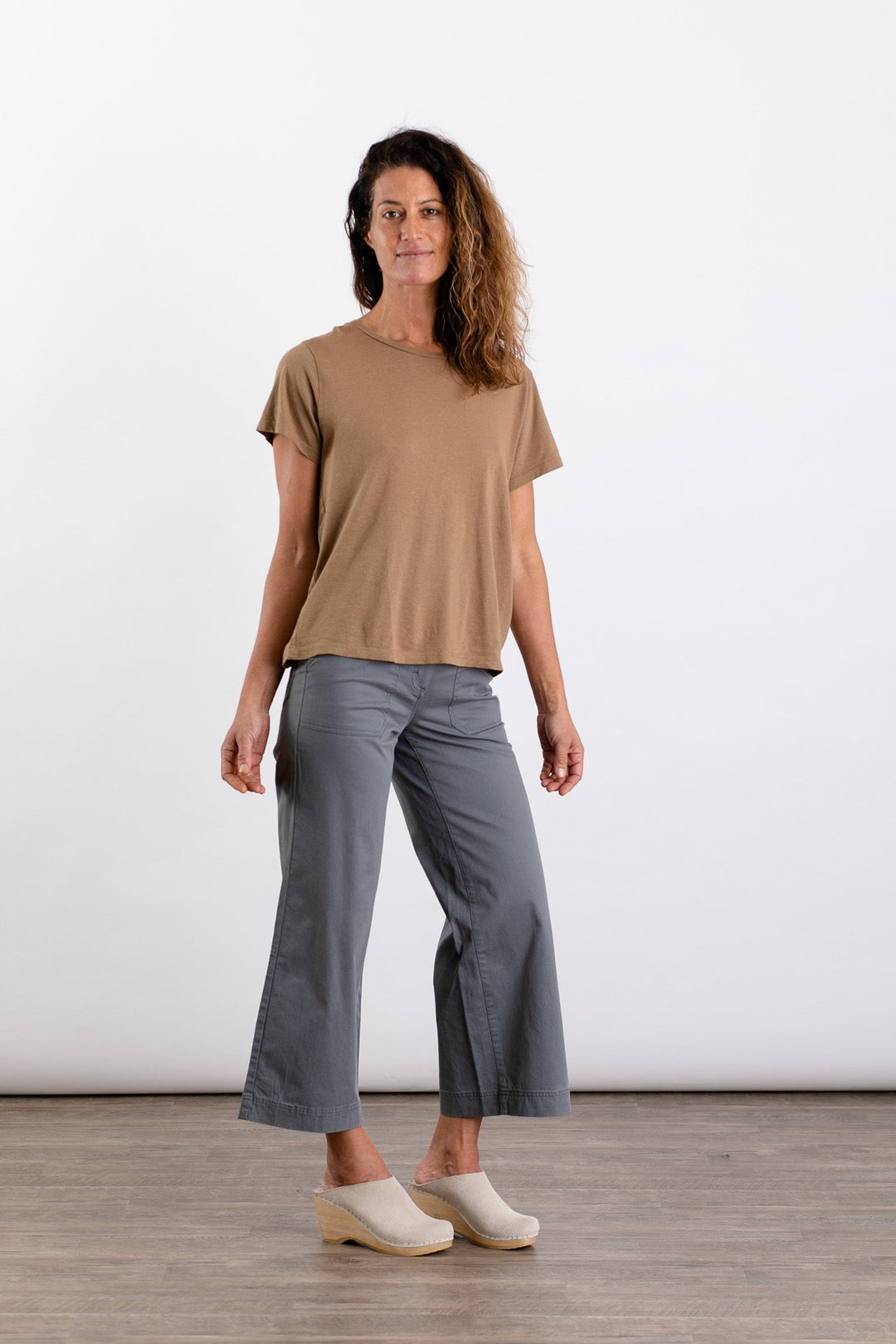 A woman with wavy brown hair wears the Jungmaven Ojai Tee in Coyote, paired with gray wide-leg pants and light clogs. She stands on a wooden floor against a white background, looking at the camera with a slight smile.