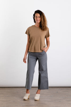 A woman stands on a wooden floor wearing the Jungmaven Ojai Tee in Coyote and gray wide-leg pants, paired with light-colored clogs. She has one hand in her pocket and smiles slightly against a plain white background.