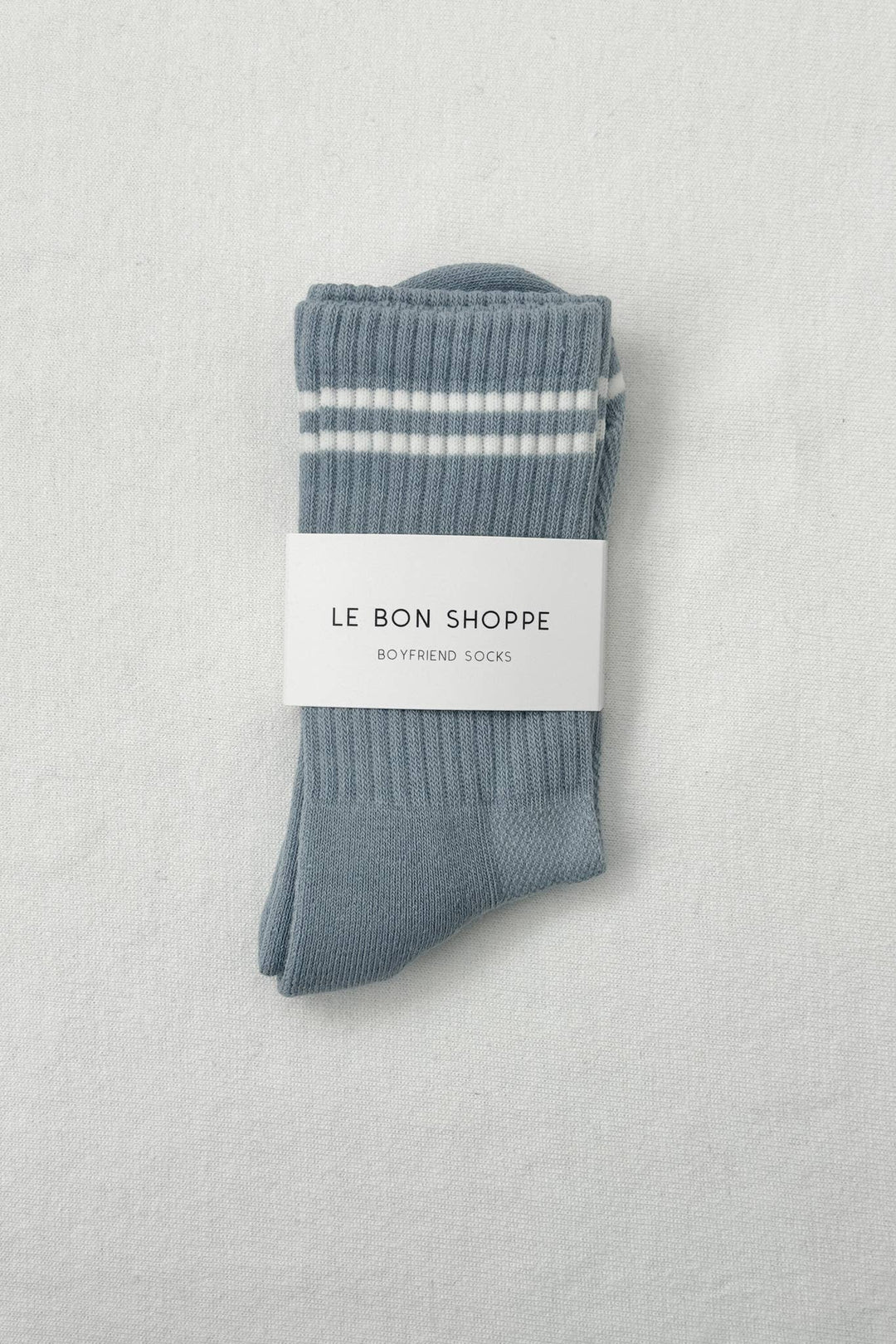 A pair of Le Bon Shoppe - Boyfriend Socks in Blue Grey with two white stripes near the top, wrapped in a branded label—an ideal cute and sporty addition to your sock collection.