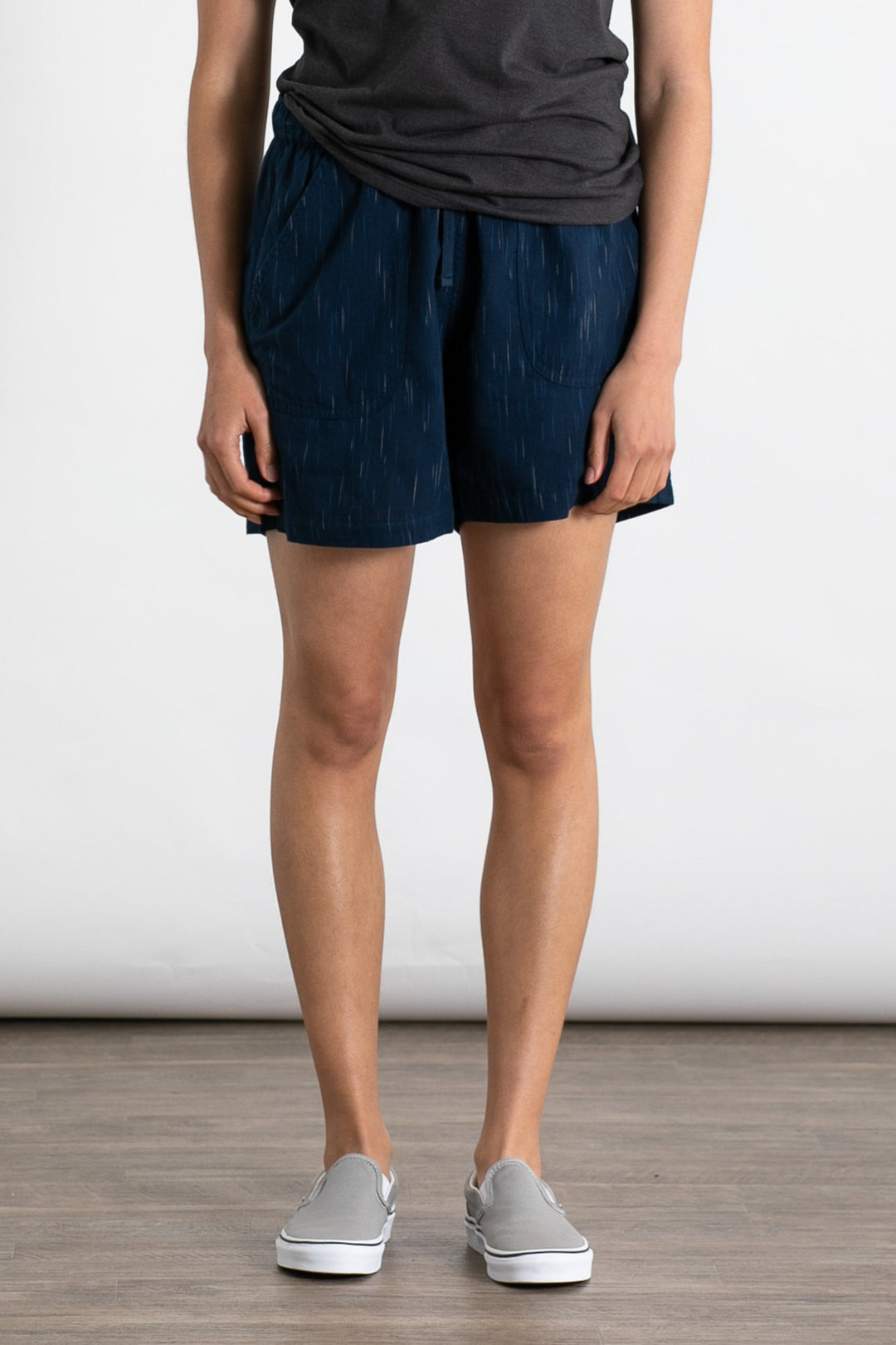 A person stands indoors on a wood floor against a plain white background, wearing Bridge & Burn’s Nico Short in Navy Space Dye with subtle vertical patterns and an elastic waist, paired with a dark gray t-shirt and gray slip-on shoes. Head not visible.
