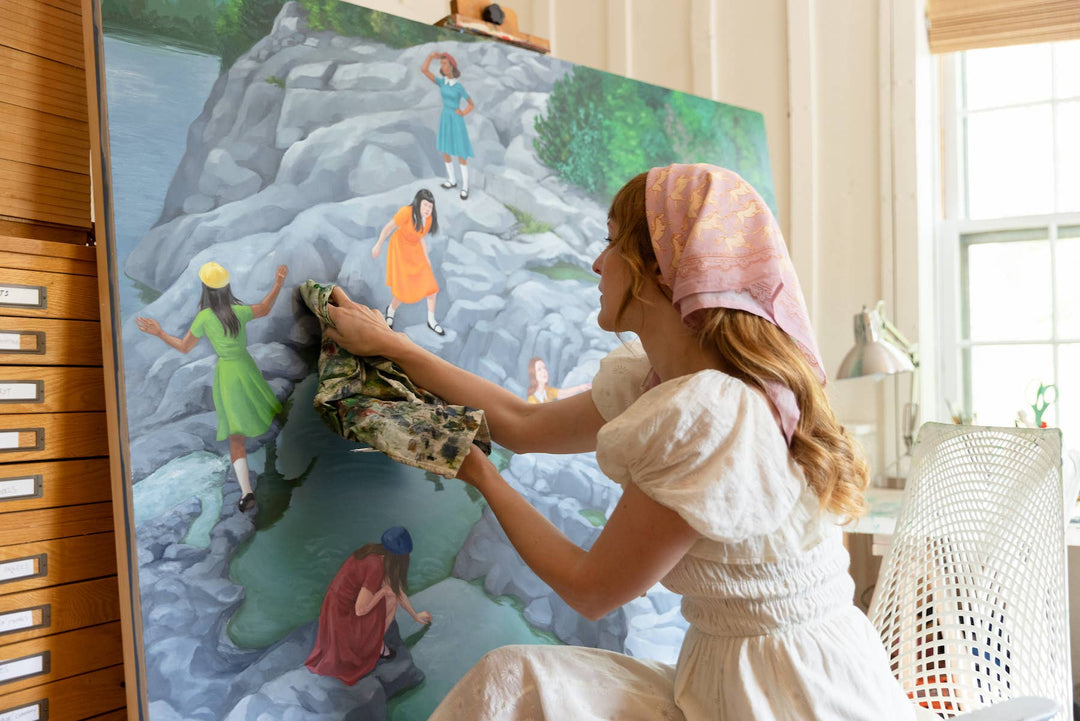 A woman wearing the Handker Bandanas 34" Wild Rag Horses Bandana sits in a bright studio, painting a large canvas of people walking on rocky terrain beside a pool of water.