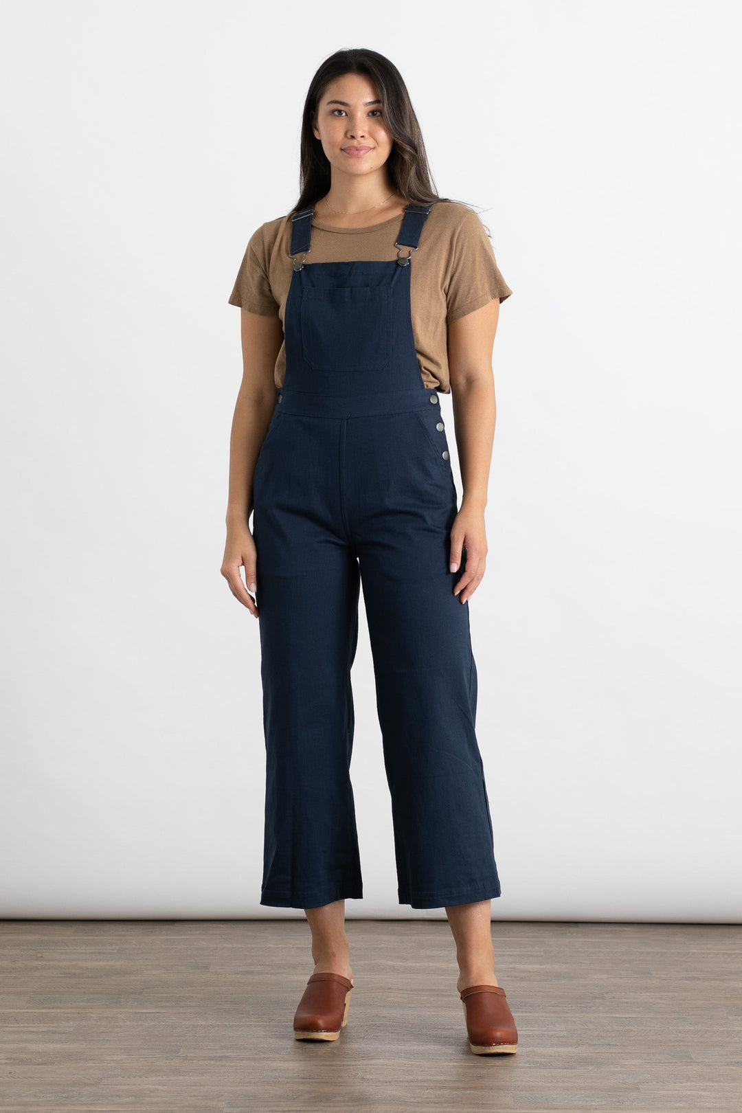 A woman with long dark hair stands on a wooden floor against a white background, wearing Bridge & Burn’s Roscoe Overall in Navy Canvas, a tan t-shirt, and brown clogs. Her arms are relaxed by her sides and she has a neutral expression.